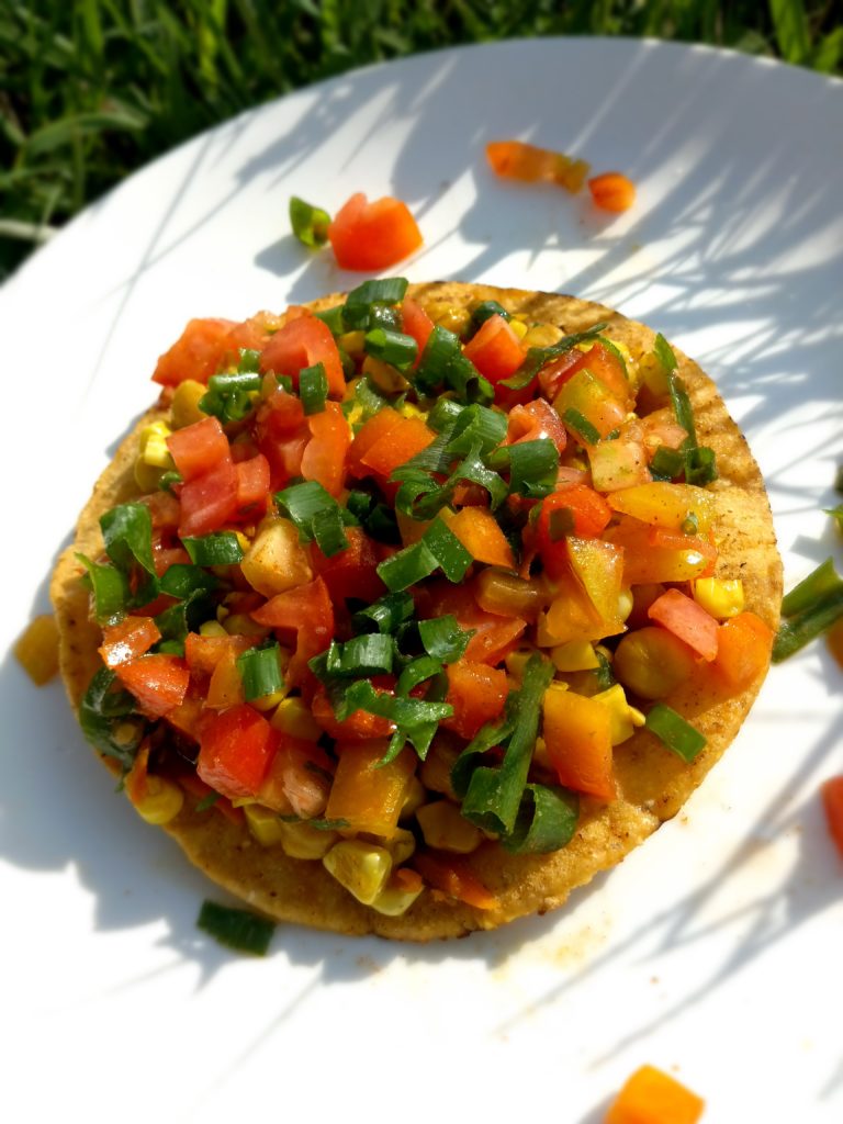 One Chickpea, Tomato, & Sweet Corn Tostada on white plate with green grass in the background.