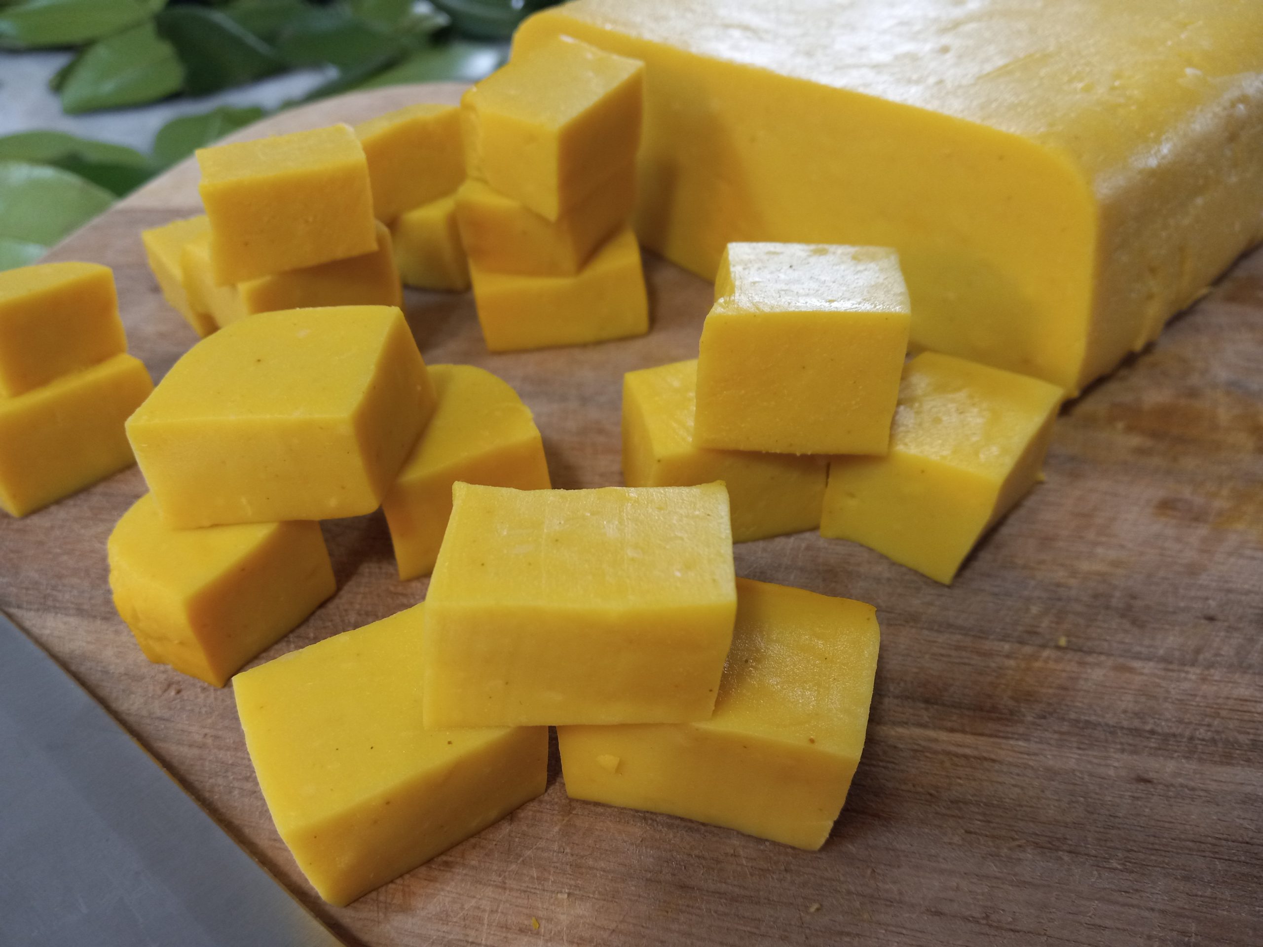 small cubes of chickpea tofu on a wooden cutting board with a big block of tofu, green leave, and a kitchen knife in the background.