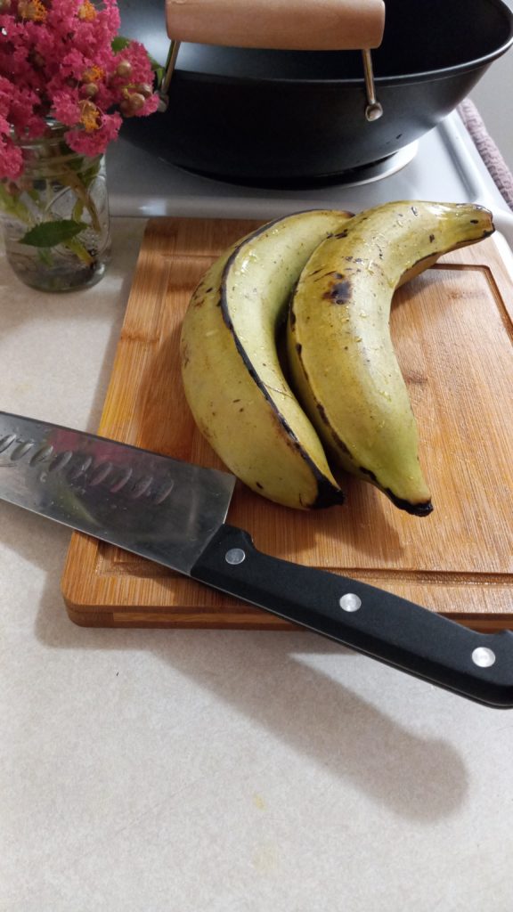 two green plantains on a wooden cutting board with a pink flower and pot in the background